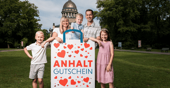 aktionsseite_anhalt gutschein_347x180 Foto von einer Familie, in der Mitte eine große Einkaufstüte mit der Aufschrift "Anhalt Gutschein" vor dem Mausoleum Dessau